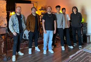 Bandfoto Aron Weise: Sechs Personen stehen nebeneinander in einem Proberaum. Sie tragen überwiegend dunkle, legere Kleidung wie T-Shirts, Hoodies und Jacken. Im Hintergrund sind Musikinstrumente und Verstärker zu sehen, der Boden ist aus Holz. Die Gruppe blickt ruhig in die Kamera, die Atmosphäre wirkt konzentriert und professionell.