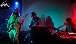 Bandfoto Chee Awai:Dreiköpfige Band auf einer dunklen Bühne bei einem Livekonzert. Links steht ein Musiker mit E-Gitarre und Mikrofon, in der Mitte spielt ein weiterer Musiker Bass, rechts sitzt ein Musiker an einem Keyboard. Blau-rote Bühnenbeleuchtung und Nebel erzeugen eine intensive Konzertatmosphäre.