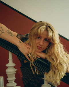 Bandfoto Kajsa Alma: Porträt einer Musikerin mit langen, welligen, blonden Haaren. Sie lehnt sich mit verschränkten Armen über ein schwarzes Treppengeländer und blickt direkt in die Kamera. Im Hintergrund sind eine rote Wand und weiße Treppenelemente zu sehen. An ihren Armen sind mehrere Tattoos sichtbar.