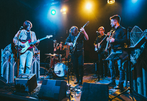 Bandfoto Holy Fanda & the Reverends: Eine fünfköpfige Band steht auf einer beleuchteten Bühne in einem Club. In der Mitte steht eine Person mit langen Haaren und Gitarre am Mikrofon, sie scheint gerade zu singen. Links und rechts spielen weitere Personen E-Gitarre und Bass, im Hintergrund sitzt eine Person am Schlagzeug. Die Bühne ist in blaues und gelbes Licht getaucht, vor der Band stehen mehrere Lautsprecher. Die Atmosphäre wirkt wie ein Live-Konzert mit energetischer Stimmung.