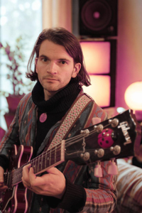 Bandfoto Harry-Hudson-Taylor: Musiker mit Gitarre im Innenraum (Porträt)
Eine Person mit schulterlangem dunklem Haar sitzt in einem Innenraum und hält eine E-Gitarre. Sie blickt ruhig in die Kamera. Die Person trägt einen gemusterten Mantel und einen dunklen Schal. Im Hintergrund stehen Lautsprecher und warmes, indirektes Licht, das eine ruhige, wohnliche Atmosphäre erzeugt.