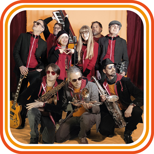 Bandfoto Hiddit: Große Bandgruppe im Studio (Gruppenfoto)
Eine große Band aus mehreren Personen posiert gemeinsam in einem Innenraum vor einem farbigen Hintergrund mit grafischem Rahmen. Die Bandmitglieder stehen und knien in mehreren Reihen und halten verschiedene Instrumente, darunter Saxofon, Trompete, Gitarre und Keyboard. Die Kleidung ist überwiegend in Rot-, Schwarz- und Grautönen gehalten. Alle Personen schauen in unterschiedliche Richtungen, die Szene wirkt lebendig und verspielt.