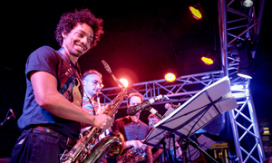 Bandfoto Noisy Neighbours Dance Party: Bläser auf Bühne bei Live-Konzert
Mehrere Musiker stehen auf einer Bühne und spielen Blasinstrumente während eines Live-Konzerts. Im Vordergrund spielt eine Person mit lockigem Haar ein Saxofon und lächelt dabei. Weitere Musiker sind dahinter zu sehen, teilweise mit Notenständern. Die Bühne ist mit buntem Bühnenlicht beleuchtet, vor allem in Rot- und Violetttönen. Die Stimmung wirkt energiegeladen und dynamisch.