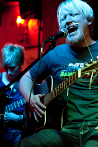 Bandfoto ge$h: Nahaufnahme eines Sängers mit sehr hellblondem Haar, der mit geschlossenen Augen intensiv in ein Mikrofon singt. Er spielt gleichzeitig eine Akustikgitarre. Sein Gesichtsausdruck wirkt emotional und kraftvoll. Im Hintergrund ist unscharf eine weitere Person mit E-Gitarre zu sehen. Das Licht ist überwiegend rot und blau, was dem Bild eine warme, intime Club-Atmosphäre verleiht.