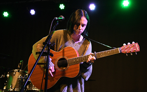 Bandfoto Mel Robin: Musikerin mit Akustikgitarre auf Bühne

Eine Person mit kurzer dunkler Frisur spielt eine Akustikgitarre auf einer Bühne. Sie steht vor einem Mikrofon. Im Hintergrund sind Schlagzeug und Bühnenlichter in Grün und Gelb zu sehen.