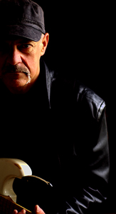 Bandfoto Wolfgang Scheele Band: Porträt eines Gitarristen

Ein Mann steht halb im Schatten vor schwarzem Hintergrund. Er trägt eine dunkle Schirmmütze und eine schwarze Lederjacke. Sein Gesicht ist nur von der linken Seite beleuchtet, wodurch ein starker Hell-Dunkel-Kontrast entsteht. Er hat einen grauen Schnurrbart und blickt ernst nach vorne. In seinen Händen hält er den Hals einer hellen E-Gitarre, die unten links im Bild zu sehen ist. Das Bild wirkt sehr stimmungsvoll und konzentriert sich auf die Person und das Instrument.