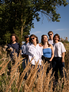 Bandfoto Eeden: Sechs Musiker stehen in einer Wiese mit hohem Gras vor Bäumen und blauem Himmel. Die Band trägt helle Sommerkleidung und blickt in verschiedene Richtungen. Die Aufnahme entstand im Freien bei Tageslicht und vermittelt eine natürliche Atmosphäre.