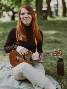 Bandfoto Lina Piacentini: Musikerin mit Ukulele im Park
Eine Musikerin sitzt auf einer Decke in einem Park und hält eine Ukulele in den Händen. Sie trägt einen dunklen Rollkragenpullover und helle Hose. Neben ihr steht eine Trinkflasche, im Hintergrund sind Bäume und eine Grünfläche zu sehen.