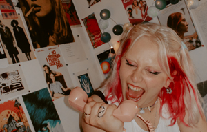 Foto Last Laugh, Blonde Musikerin schreit in Telefon Hörer, im Hintergrund Wand mit Fotos
