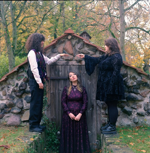 Bandfoto Eibhlin: Drei Musikerinnen vor steinerner Hütte
Drei Musikerinnen posieren vor einer kleinen steinernen Hütte in einem herbstlichen Park. Eine Musikerin sitzt vor der Tür, während die beiden anderen auf niedrigen Mauern stehen. Die Band trägt dunkle Kleidung, die Aufnahme wirkt wie ein inszeniertes Gruppenporträt.