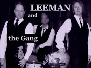 Foto der Band Leeman and The Gang.Drei Männer in weißen Hemden, dunklen Westen und Krawatten stehen vor dunklem Hintergrund. Zwei von ihnen halten Instrumente: ein Bass und eine Gitarre, während der dritte hinter einem Schlagzeug sitzt. Alle blicken ernst in die Kamera. In großer, heller Schrift steht „LEEMAN and the Gang“ über dem Bild. Die Aufnahme ist in Schwarz-Weiß mit leicht violettem Ton gehalten und wirkt klassisch und stilvoll.