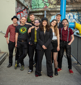 Foto der Band Arte Bella & The Fellas. Eine Gruppe von acht Personen steht auf einem urbanen Gehweg mit Graffiti-Wänden im Hintergrund. Alle Personen schauen freundlich in die Kamera. Die Gruppe ist gemischt, mit Männern und Frauen, alle in lockerer, individueller Kleidung. Eine Frau in der Mitte trägt einen dunklen Nadelstreifenanzug und eine Schirmmütze. Die anderen tragen T-Shirts, Hemden, Hüte oder Mützen. Im Hintergrund ist eine Rutsche und eine Treppe zu sehen. Es herrscht eine entspannte, kreative Atmosphäre.