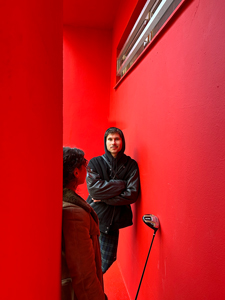 Band Foto Karl Rich. Zwei Personen stehen in einem sehr schmalen, leuchtend roten Gang. Wände, Decke und Boden sind komplett in kräftigem Rot gestrichen, was eine intensive Atmosphäre erzeugt. Eine Person lehnt mit verschränkten Armen an der rechten Wand und trägt eine schwarze Kapuzenjacke. Sie schaut ruhig und direkt in die Kamera. Die zweite Person, nur von hinten sichtbar, trägt eine braune Jacke und schaut zur ersten Person. Zwischen ihnen steht ein Golfschläger mit dem Schlägerkopf nach oben an die Wand gelehnt. Oben an der Wand befindet sich ein schmales, waagerechtes Fenster, das etwas Tageslicht hereinlässt. Die Szene wirkt durch Farbe und Perspektive sehr markant und künstlerisch.