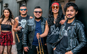Foto der Band The Klaxon. Fünf Bandmitglieder posieren lässig vor einer schwarzen Wand. Zwei Männer in Lederjacken und Sonnenbrillen halten Trompeten. Eine Frau mit langen dunklen Haaren trägt ein rotes Stirnband und ein T-Shirt mit dem Bandnamen "The Klaxon". Alle wirken selbstbewusst und freundlich. Die Kleidung ist rockig, mit viel Schwarz und Leder, was einen rebellischen Stil vermittelt. Die Gruppe steht eng beisammen und schaut direkt in die Kamera.