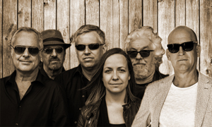 Foto der Band Midlife. Sechs Personen – fünf Männer und eine Frau – stehen in zwei Reihen vor einer Holzwand. Alle tragen Sonnenbrillen und dunkle Kleidung, bis auf einen Mann rechts in einem hellen Sakko. Die Bildstimmung ist entspannt und einheitlich, durch den Sepiafilter wirkt es warm und retroartig. Die Personen blicken gelassen in die Kamera.