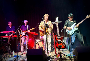 Foto der Syndicate Band. Fünf Musiker befinden sich auf einer Bühne mit schwarzem Vorhang im Hintergrund. Sie spielen ihre Instrumente: E-Gitarre, Bass, Keyboard, Schlagzeug und eine zusätzliche Gitarre steht auf einem Ständer vorne. Der Sänger in der Mitte trägt ein hell gemustertes Hemd und singt in ein Mikrofon. Die Band ist in Aktion, in eine Performance vertieft, mit buntem Bühnenlicht und Kabeln auf dem Boden. Die Stimmung wirkt energiegeladen und musikalisch fokussiert.