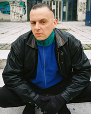 Bandfoto Kalaszinco: Ein Mann sitzt in einer urbanen Umgebung mit grauen Pflastersteinen und Graffiti im Hintergrund. Er trägt eine schwarze Lederjacke über einem blauen Pullover mit grünem Kragen. Seine Hände sind in schwarzen Handschuhen gefaltet, er schaut ernst und direkt in die Kamera. Die Körperhaltung ist selbstbewusst, fast herausfordernd.