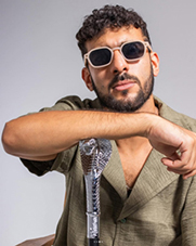 Bandfoto Nista: Ein Mann mit lockigem, kurzem Haar und Bart posiert vor neutralem grauen Hintergrund. Er trägt ein olivgrünes Hemd, weiße Sonnenbrille und stützt den rechten Arm auf ein glitzernd verziertes Mikrofon. Sein Blick geht selbstbewusst in die Kamera, die Pose wirkt cool und lässig.