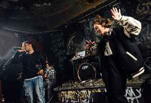 Bandfoto Música pa' pobre

Auf einer kleinen Bühne mit Graffiti-Hintergrund treten zwei Musiker*innen auf. Links steht ein Mann mit schwarzem Oberteil und heller Jeans, er hält ein Mikrofon und singt. Rechts daneben steht eine Frau in weiter schwarzer Kleidung und heller Collegejacke. Sie bewegt sich mit erhobenen Händen und wirkt tänzerisch, während sie ins Publikum schaut. Hinter ihnen sieht man ein Schlagzeug, Verstärker und weitere Bandmitglieder. Die Stimmung ist energiegeladen und urban.