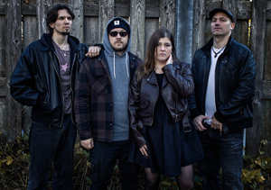Bandfoto Rising Alma: Vier Personen stehen vor einem alten Holzzaun. Zwei Männer tragen dunkle Jacken und Mützen, eine Frau in der Mitte trägt ein schwarzes Kleid und eine Lederjacke. Die Stimmung wirkt ernst und konzentriert, das Licht ist kühl und herbstlich.