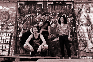 Bandfoto Plague Pets: Fünf junge Erwachsene posieren vor einer Wand mit großflächiger Graffiti-Kunst. Eine Person hockt vorn, die anderen stehen dahinter. Alle wirken entspannt und gut gelaunt. Das Foto ist in warmen Sepia-Tönen gehalten und vermittelt eine alternative, urbane Atmosphäre.