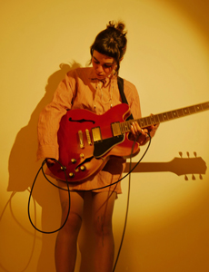 Band Foto Vasylysa:

Eine Musikerin steht vor einer gelb beleuchteten Wand und spielt eine rote E-Gitarre. Sie trägt ein langes, hellbraunes Hemd und blickt konzentriert nach unten auf ihr Instrument. Das Licht wirft einen weichen Schatten an die Wand, wodurch das Bild eine warme, intime Atmosphäre bekommt.