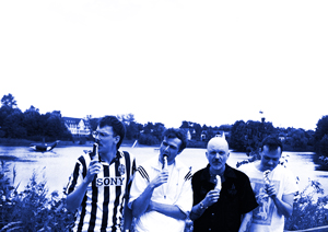 Bandfoto Samt Martin: Vier Musiker am See in Schwarzweiß
Vier Musiker stehen nebeneinander am Ufer eines Sees und halten Getränke in den Händen. Sie tragen unterschiedliche Freizeitkleidung, darunter ein gestreiftes Fußballtrikot und helle Hemden. Im Hintergrund sind Wasser, Bäume und Häuser zu sehen. Die Aufnahme ist in kontrastreichen Blau- und Weißtönen gehalten und vermittelt eine lockere, sommerliche Atmosphäre.