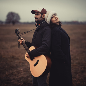 Bandfoto The Jingle Jangle Morning:
Zwei Musiker stehen Rücken an Rücken auf einem weiten, braunen Feld unter bewölktem Himmel. Der Musiker links trägt eine Baseballkappe, einen dunklen Mantel und hält eine akustische Gitarre. Die Musikerin rechts trägt einen langen schwarzen Mantel und eine helle Fellmütze. Beide blicken in unterschiedliche Richtungen.