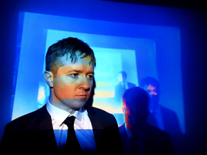 Bandfoto deadPEASNTS: Musiker im Anzug vor blauer Lichtprojektion
Ein Musiker mit kurzen Haaren trägt einen schwarzen Anzug, ein weißes Hemd und eine schwarze Krawatte. Er steht vor einer großflächigen blauen Lichtprojektion mit ineinander verschachtelten Rechtecken und blickt nachdenklich zur Seite. Im Hintergrund sind schemenhaft weitere Musiker zu erkennen. Die Aufnahme wirkt geheimnisvoll und atmosphärisch.