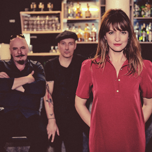 Bandfoto Paule und Band - Christmas Konzert: Die Schauspielerin Ina Paule Klink im roten Kleid steht im Vordergrund einer Bar und blickt direkt in die Kamera. Hinter ihr sitzen zwei Männer auf Barhockern; beide tragen dunkle Kleidung, einer mit Sonnenbrille oben auf dem Kopf, der andere mit Mütze. Die Stimmung wirkt warm und intim.