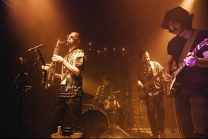 Bandfoto Cabin Crew: Vier Musiker spielen live auf einer Clubbühne. Links steht ein Gitarrist am Mikrofon, daneben ein Saxophonist, der sein Instrument spielt. Im Hintergrund sitzt ein Schlagzeuger am Drumset. Rechts steht ein weiterer Gitarrist. Die Bühne ist in warmes, gelbliches Licht und leichten Nebel getaucht, wodurch eine dichte Konzertatmosphäre entsteht.