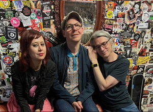 Bandfoto Front Porch Beatnik: Drei Personen sitzen vor einer bunt beklebten Wand voller Aufkleber, Poster und Grafiken. Links sitzt eine Frau mit rotem Haar, in der Mitte ein Mann mit Brille und Basecap, rechts ein weiterer Mann mit Brille und langen grauen Haaren, der seinen Kopf an die Schulter des Mannes in der Mitte lehnt. Die Stimmung wirkt entspannt und freundschaftlich.