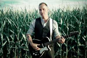 Bandfoto Wayne Jackson: Ein Mann steht in einem hohen Maisfeld und spielt eine E-Gitarre. Er trägt eine Weste über einem Hemd und hat Tätowierungen an beiden Armen. Der Himmel im Hintergrund ist hell und leicht bewölkt. Die Szene wirkt ruhig und atmosphärisch. 